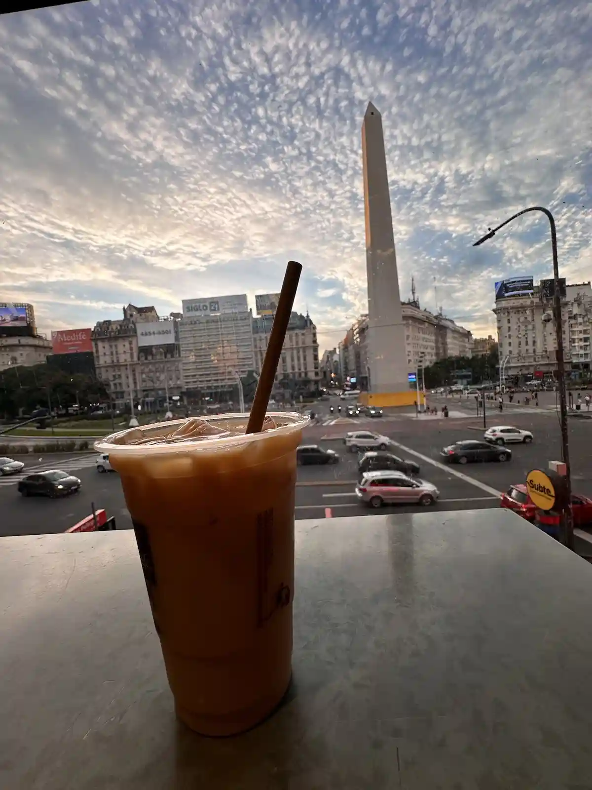 A plastic cup of iced coffee with a brown straw sits on a metal table, with a view of the Obelisco de Buenos Aires and city…