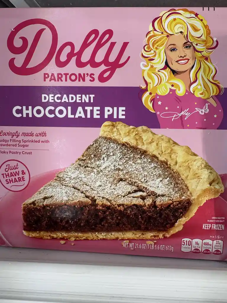 A close-up of a frozen pie box featuring a cartoon illustration of Dolly Parton with blonde hair and a pink outfit. The box…