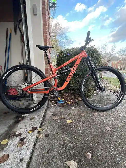 An orange Canyon mountain bike with black tires and blue pedals is parked on a concrete driveway next to a brick house…