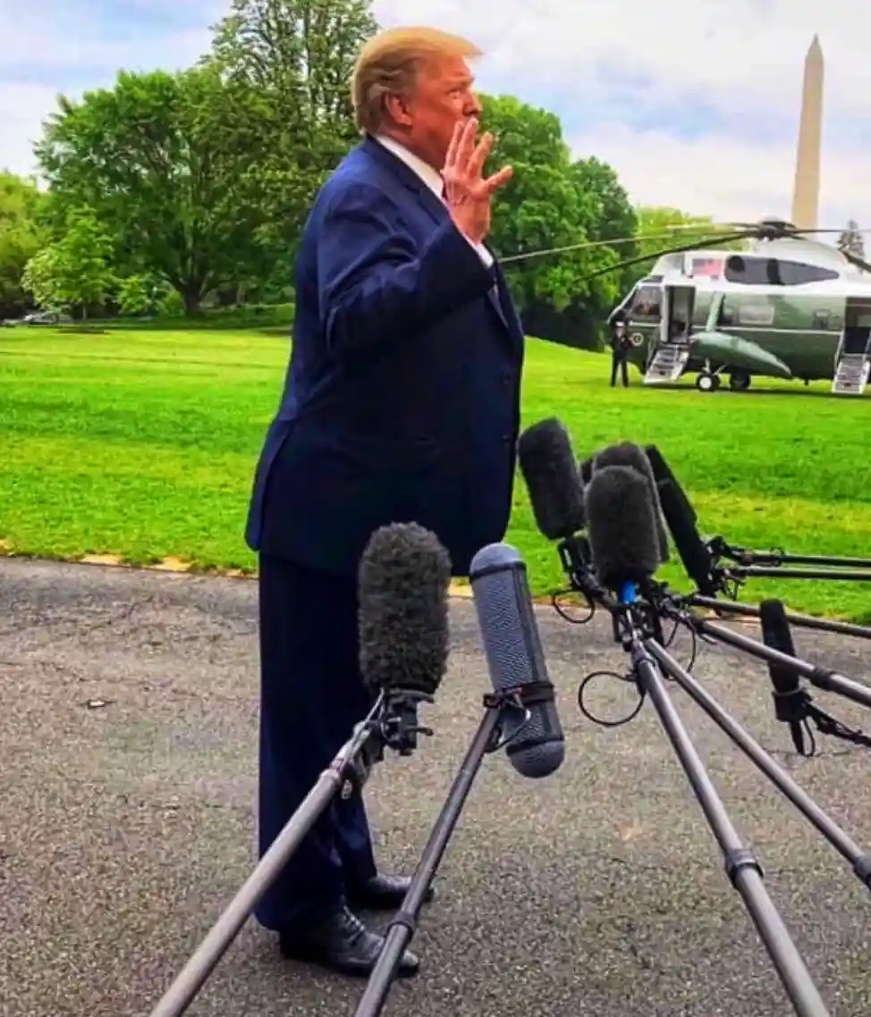 A man in a dark suit stands outdoors near microphones on stands, gesturing with his hand as if speaking. In the background…