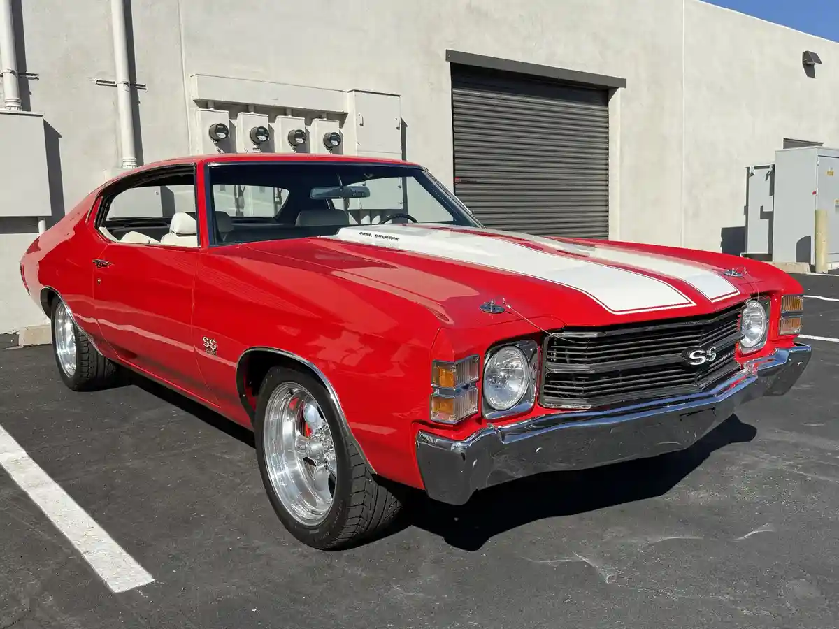 A shiny red classic Chevrolet Chevelle SS muscle car with white racing stripes is parked in a lot. The car has chrome…