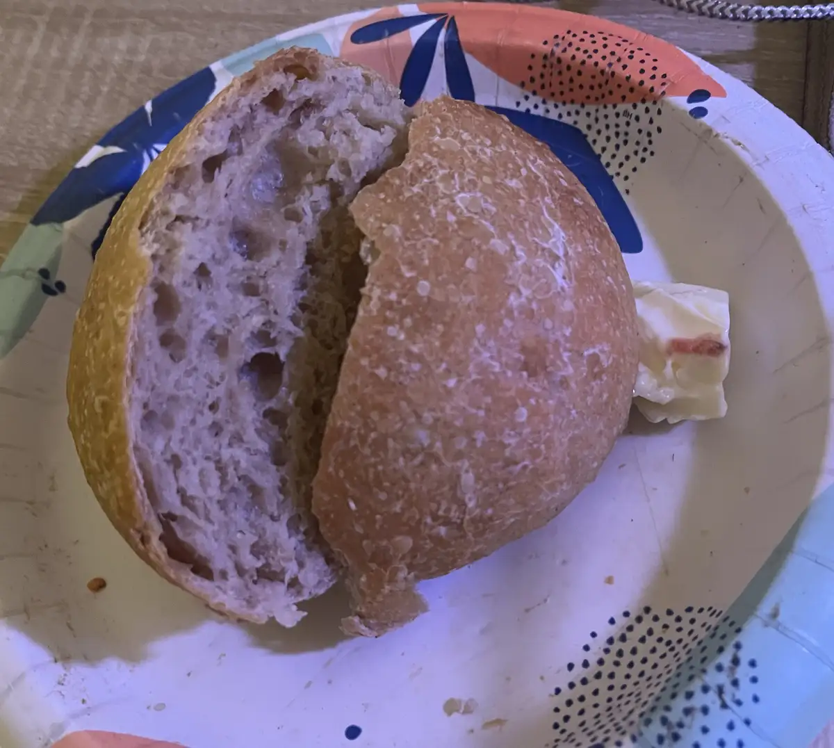 A close-up photo shows a rustic, crusty bread roll split open on a colorful paper plate, with a small pat of butter beside…