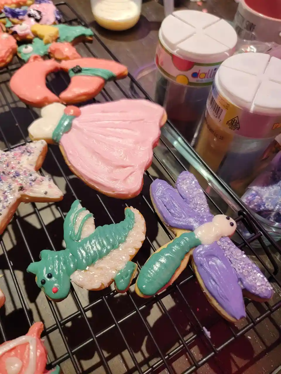 A close-up photo shows several intricately decorated sugar cookies on a black wire cooling rack. The cookies are shaped like…