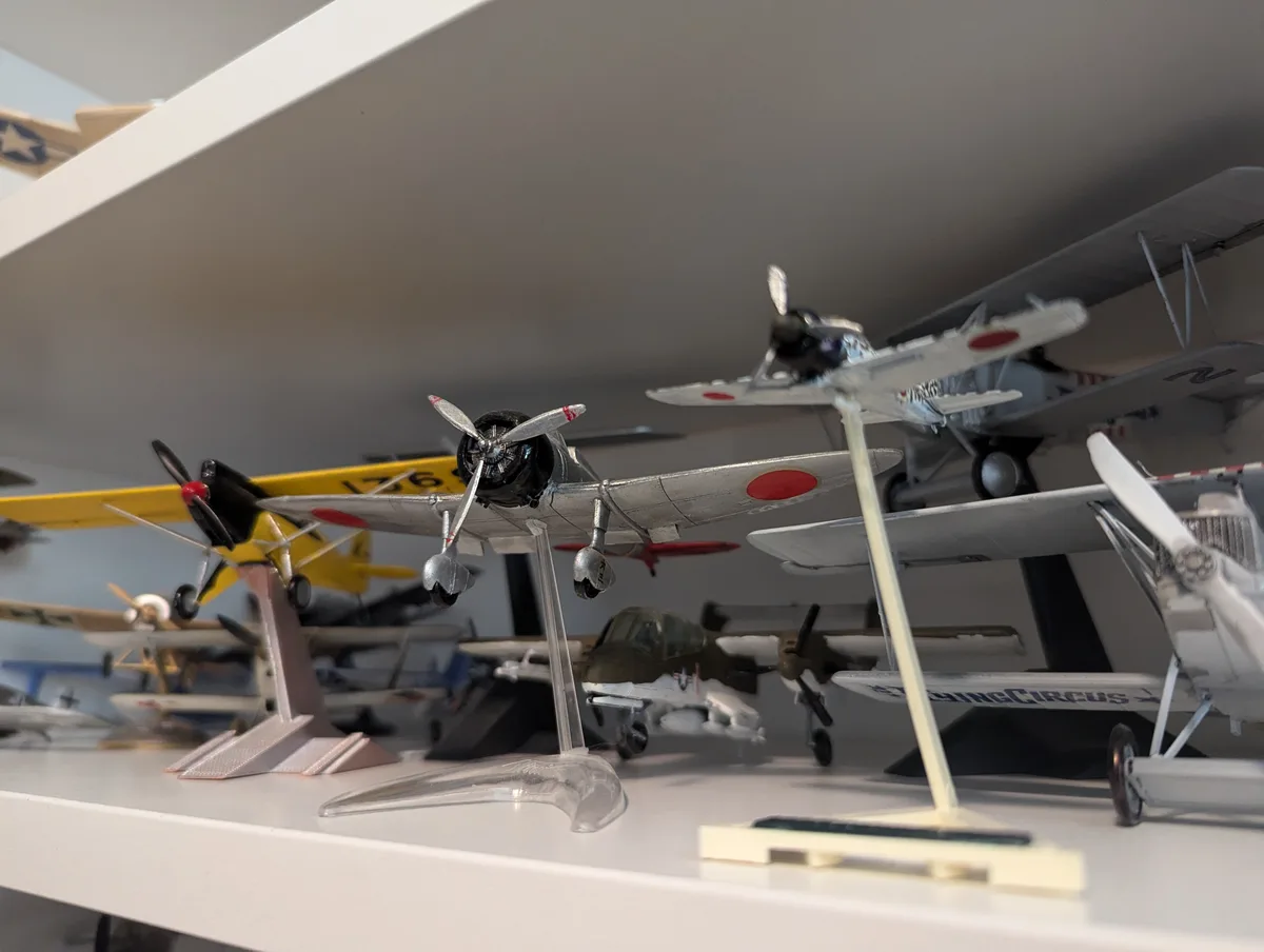 A close-up photo shows several detailed model airplanes displayed on white shelves, including a silver biplane with a red…