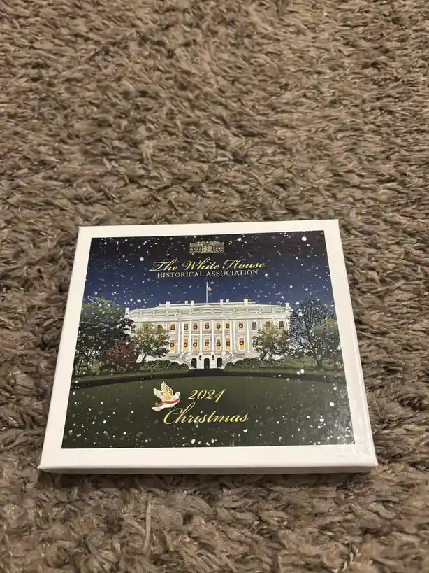 A square box with a festive illustration of the White House at night under falling snow, decorated with wreaths in its…