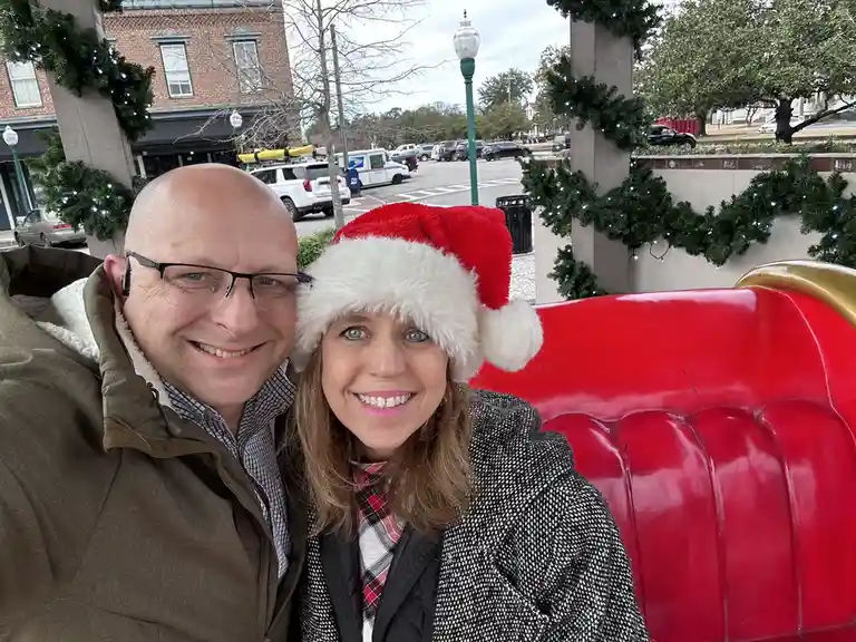 A smiling couple poses for a selfie outdoors near a red sleigh-like bench decorated with Christmas garlands. The woman wears…