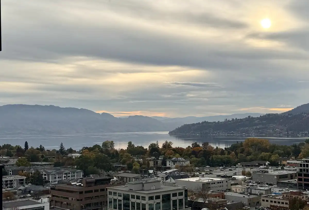 A scenic view of a city with a large lake and mountains under a cloudy sky with the sun visible. Buildings and trees with…
