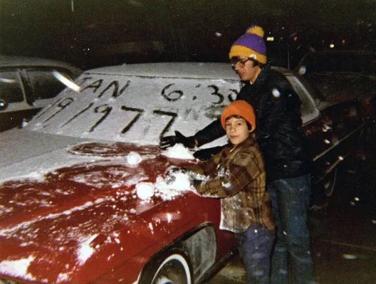 Two people, an adult and a child, are cleaning snow off a red car at night. The windshield has "FAN 6:30 19/1977" written in…