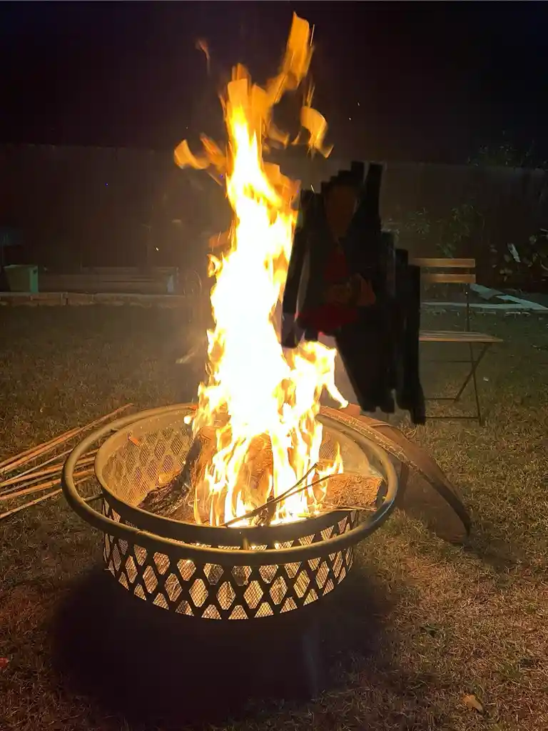 A person sits in a backyard at night beside a large, roaring fire in a metal fire pit, holding a drink. The flames are tall…