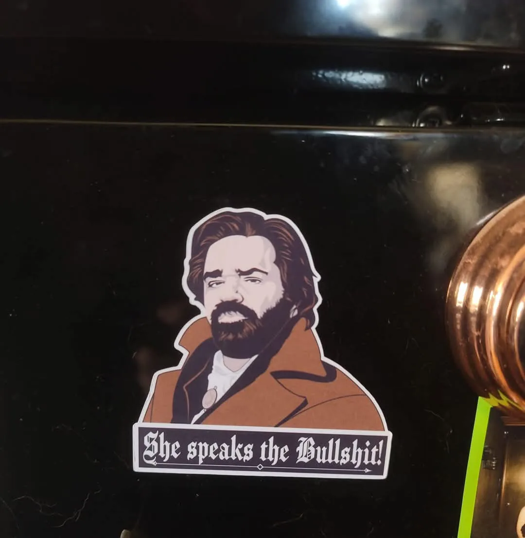 A sticker featuring a cartoon illustration of a man with a beard and brown coat, placed on a dark surface. The sticker has a…