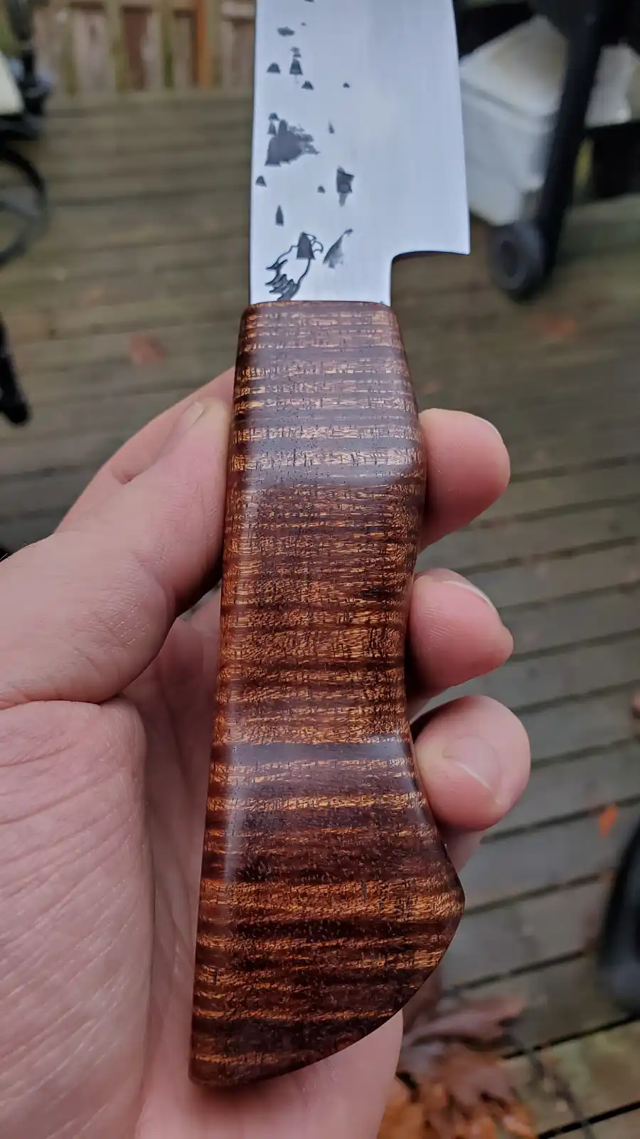 A hand holds a custom knife with a polished, dark wood handle featuring a swirling grain pattern. The blade is stainless…