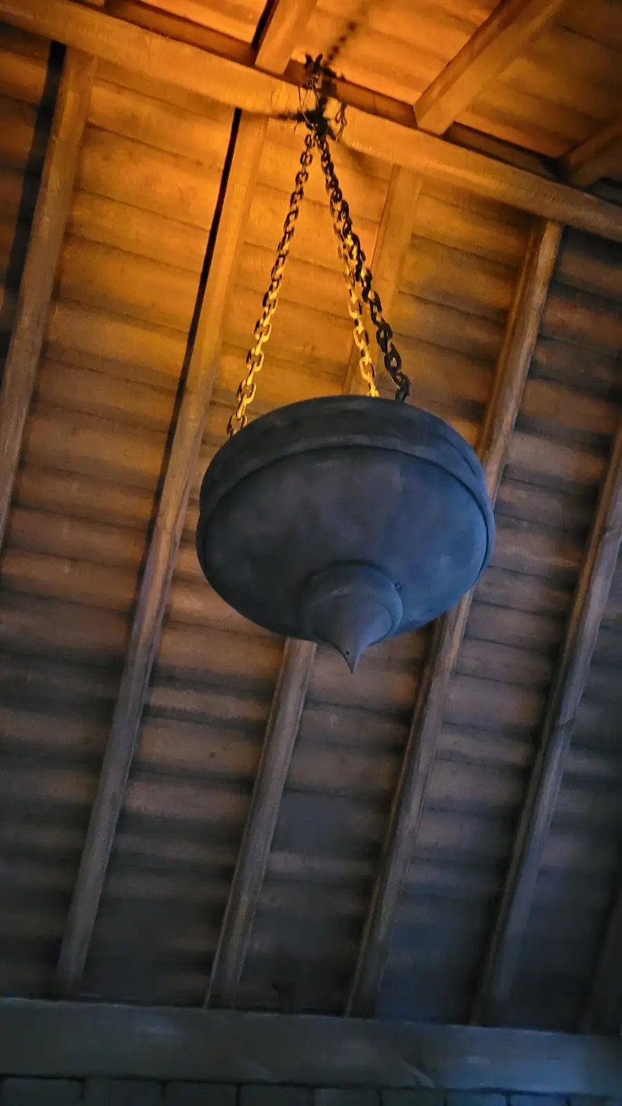 A large, dark, bell-shaped object hangs from chains against a wooden ceiling with exposed beams. The overlaid text reads…