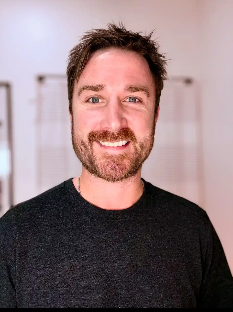A man with short brown hair and a beard smiles at the camera while wearing a black t-shirt and a silver chain. The overlaid…