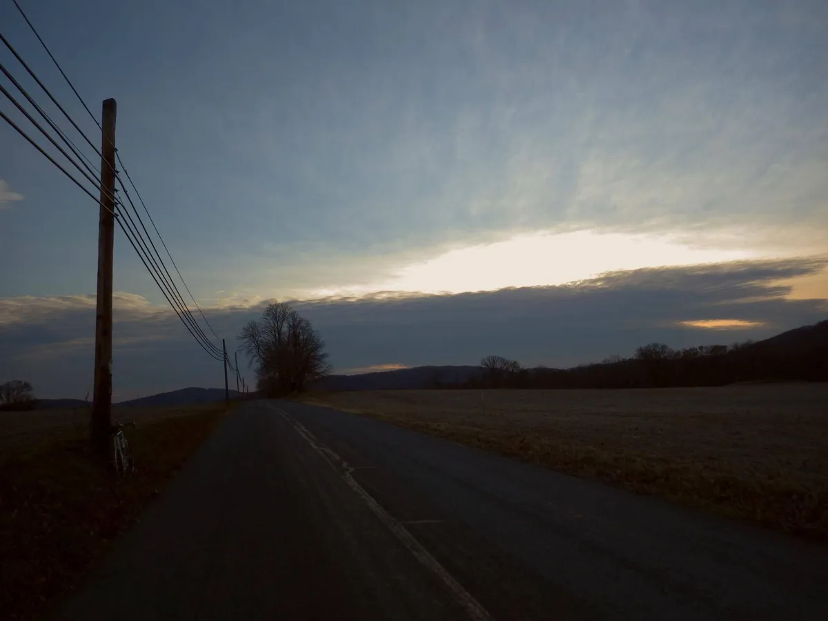 A dimly lit rural road at dusk with a bicycle leaning against a utility pole on the left. The sky shows a bright sunlit…
