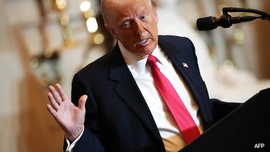 a man in a dark suit and red tie stands at a podium with a microphone, gesturing with his right hand while speaking. The…