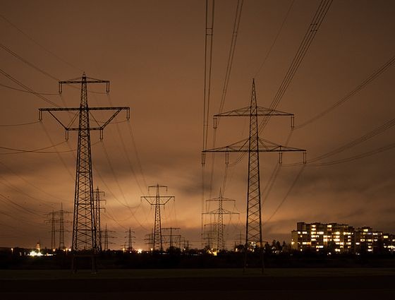 A dark, atmospheric photo of multiple electricity pylons silhouetted against a dusky orange sky, with distant city lights…