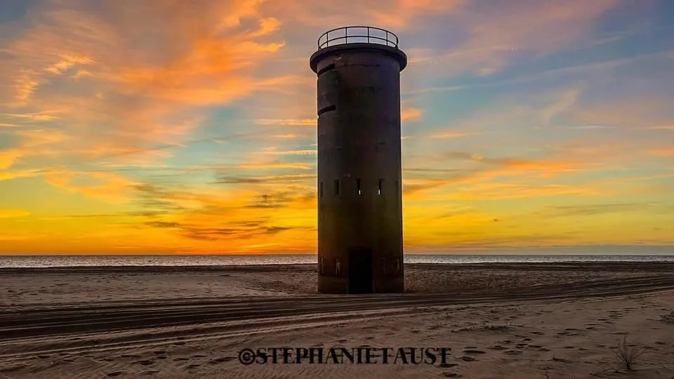 A weathered, cylindrical concrete tower stands alone on a sandy beach at sunset, with vibrant orange, yellow, and blue…