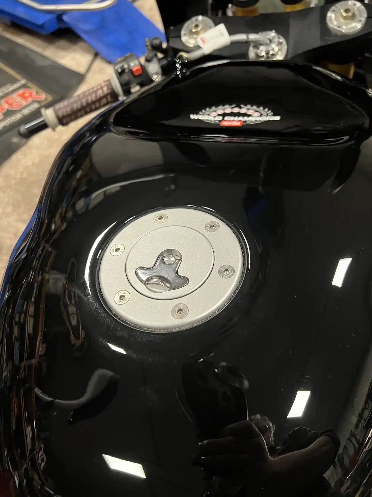 A close-up photo of a glossy black motorcycle fuel tank with a silver fuel cap and 'world champions' logo. The tank's…