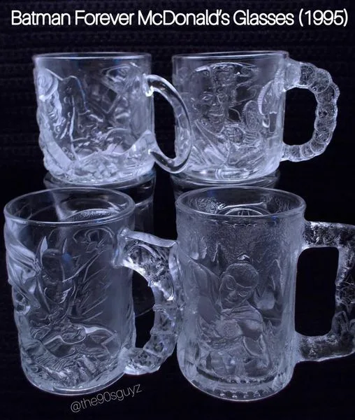 Four clear glass mugs with handles are arranged in a square on a dark surface. Each mug has an embossed scene from the 1995…