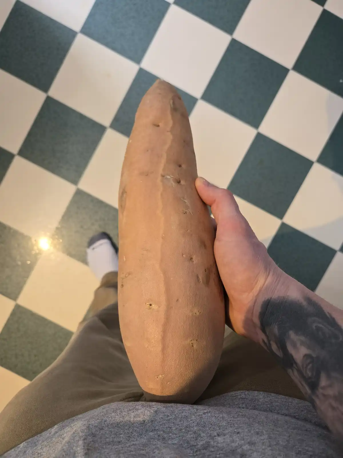 A person holds a very large, elongated sweet potato in their hand, which dominates the frame. The background is a green and…