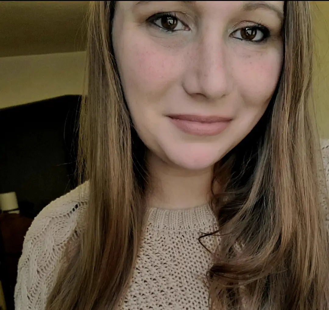A close-up selfie of a young woman with long brown hair and freckles, wearing a beige knit sweater, smiling slightly with a…