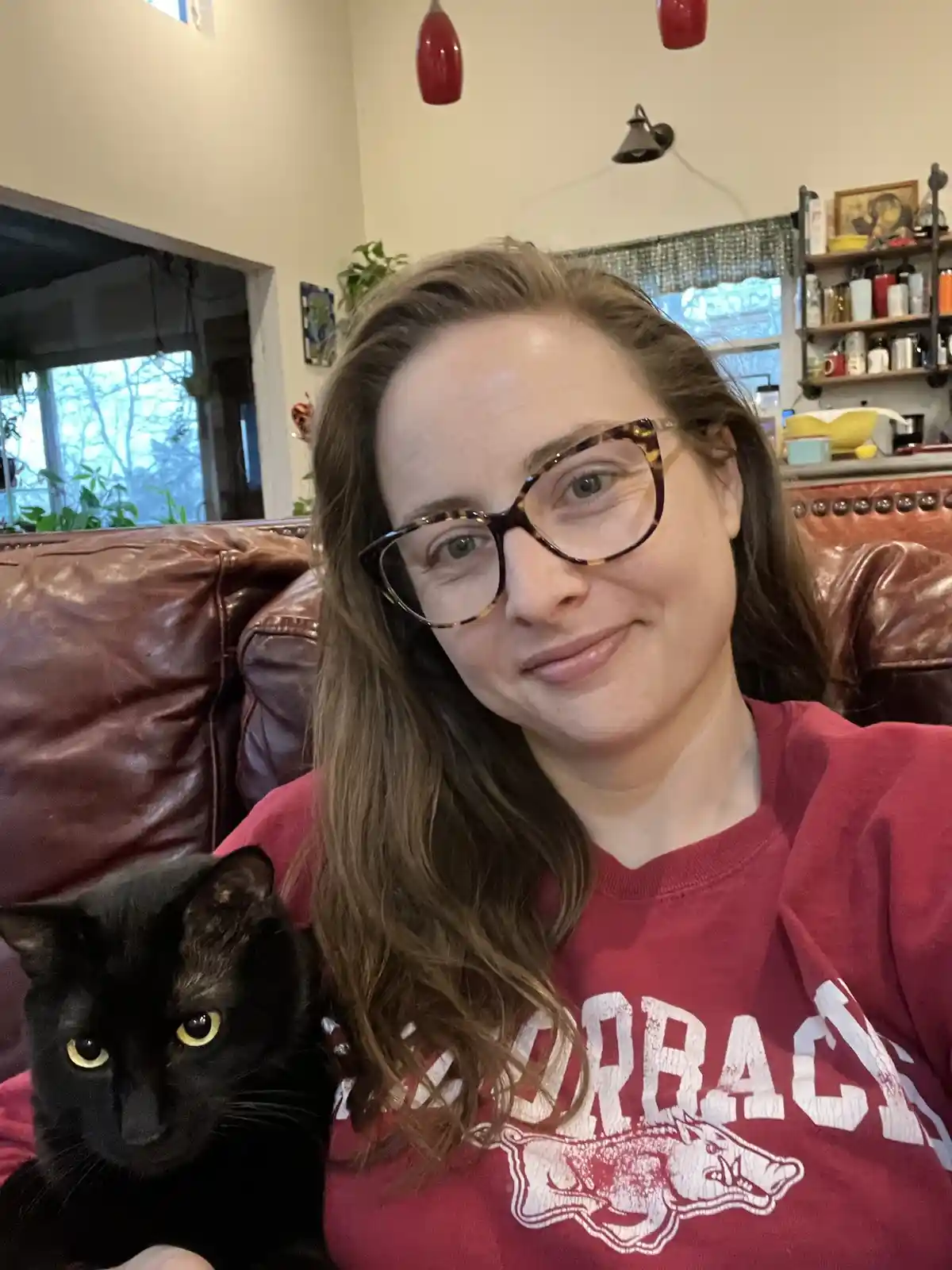A woman with glasses and a red shirt sits on a brown leather couch holding a black cat. The cat has bright yellow eyes and…