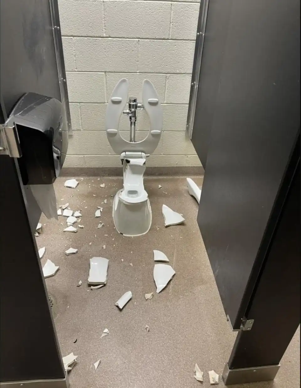 A broken toilet in a public restroom stall, with shattered porcelain pieces scattered across the floor and the toilet seat…