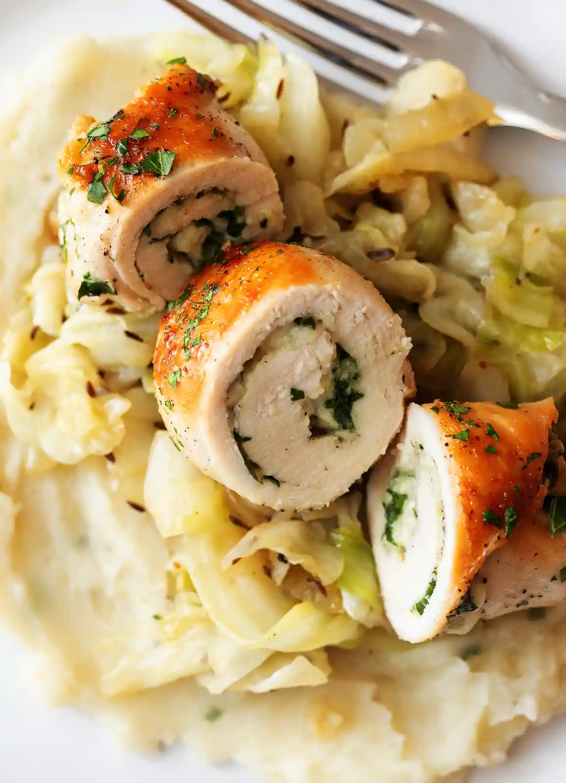 A close-up photo of a plated meal featuring sliced chicken rolls with a green herb filling, served atop sautéed cabbage and…