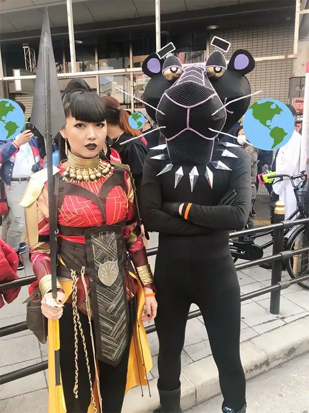 Two people in cosplay stand side by side outdoors; one is dressed as a warrior with a spear and ornate armor, the other…