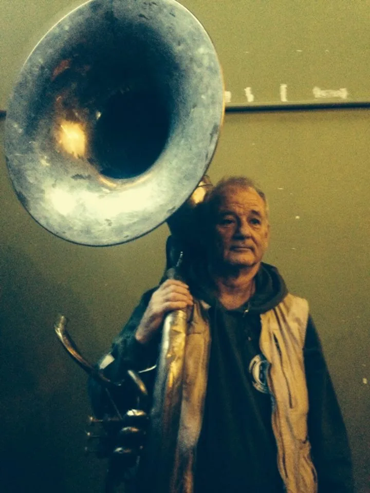 An older man with a weathered face and casual clothing holds a large, worn tuba, its bell dominating the upper half of the…