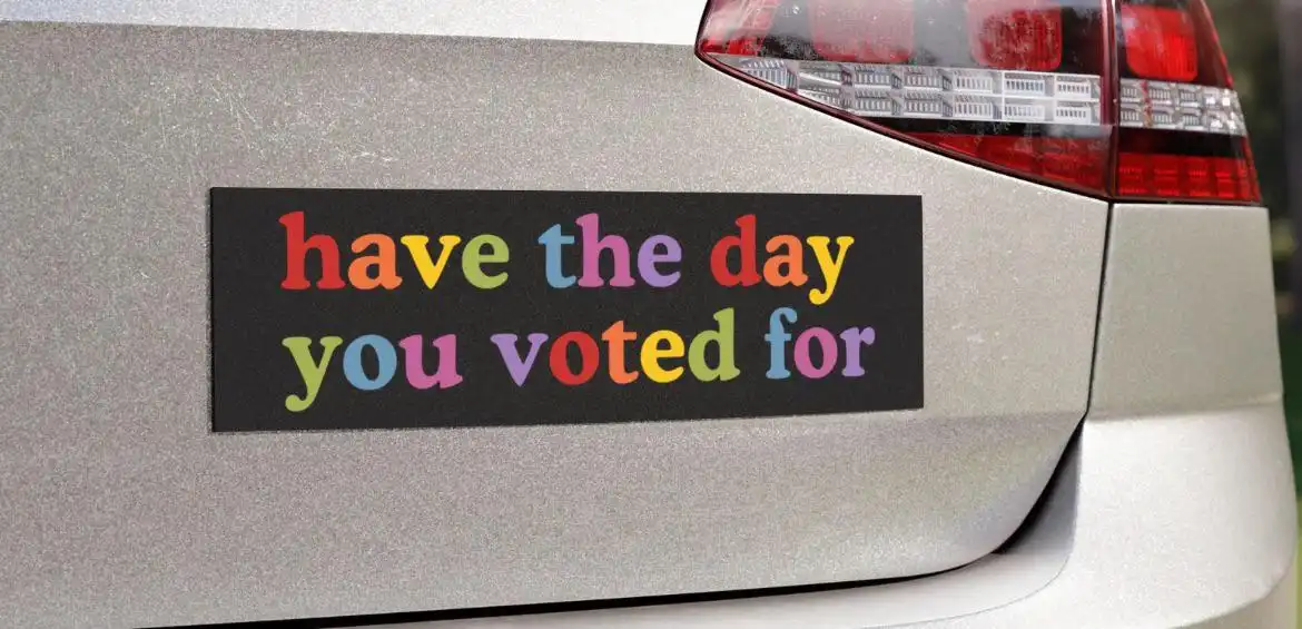 A close-up of a silver car's rear bumper featuring a black rectangular sticker with colorful, rainbow-hued text that reads…