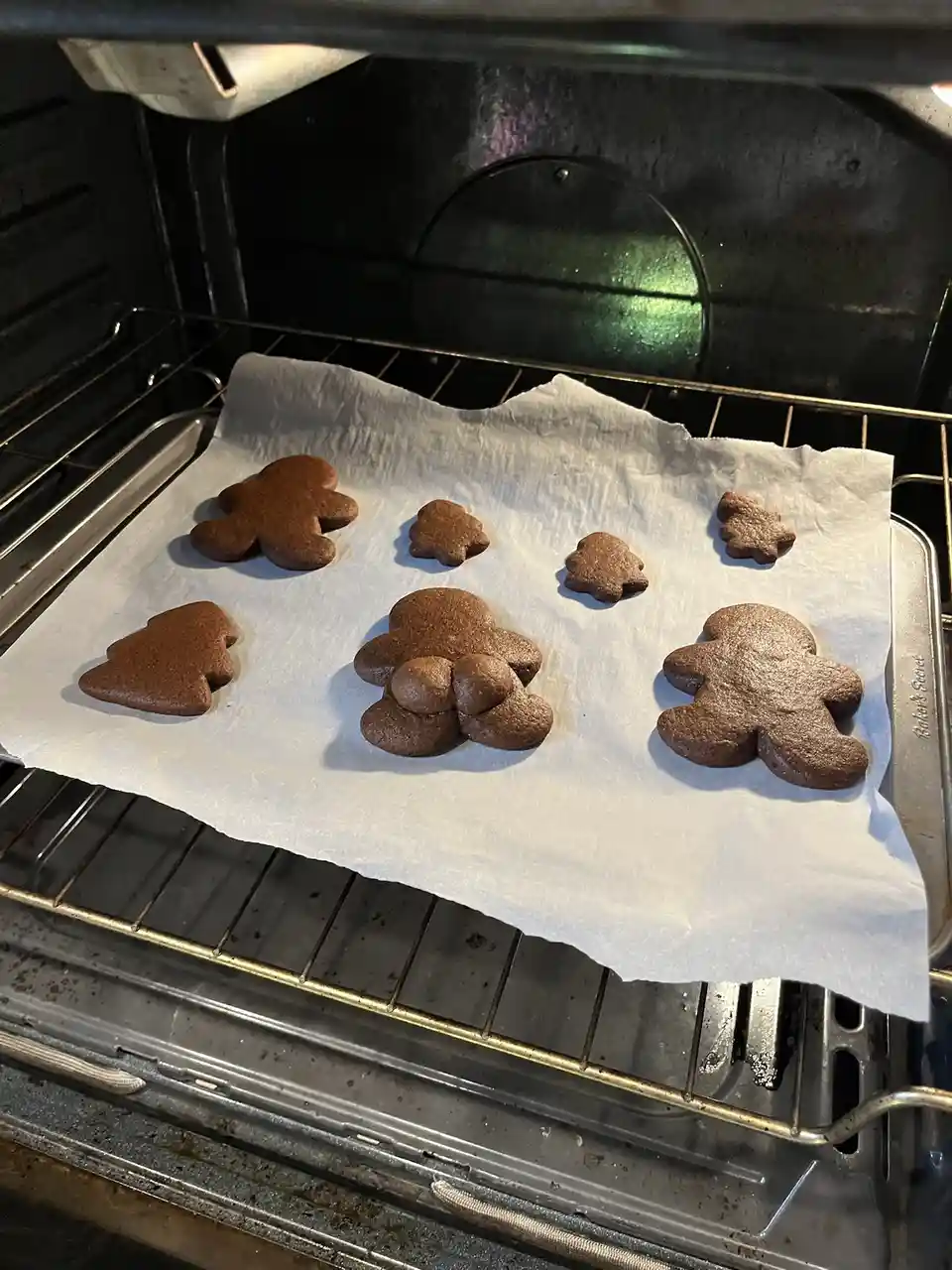 A baking sheet inside an oven holds several gingerbread cookies, including gingerbread men and trees, some of which are dark…