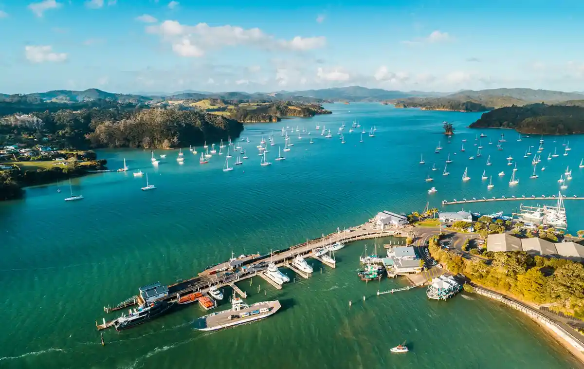 An aerial photo shows a scenic harbor filled with numerous sailboats and yachts docked at piers, surrounded by green hills…