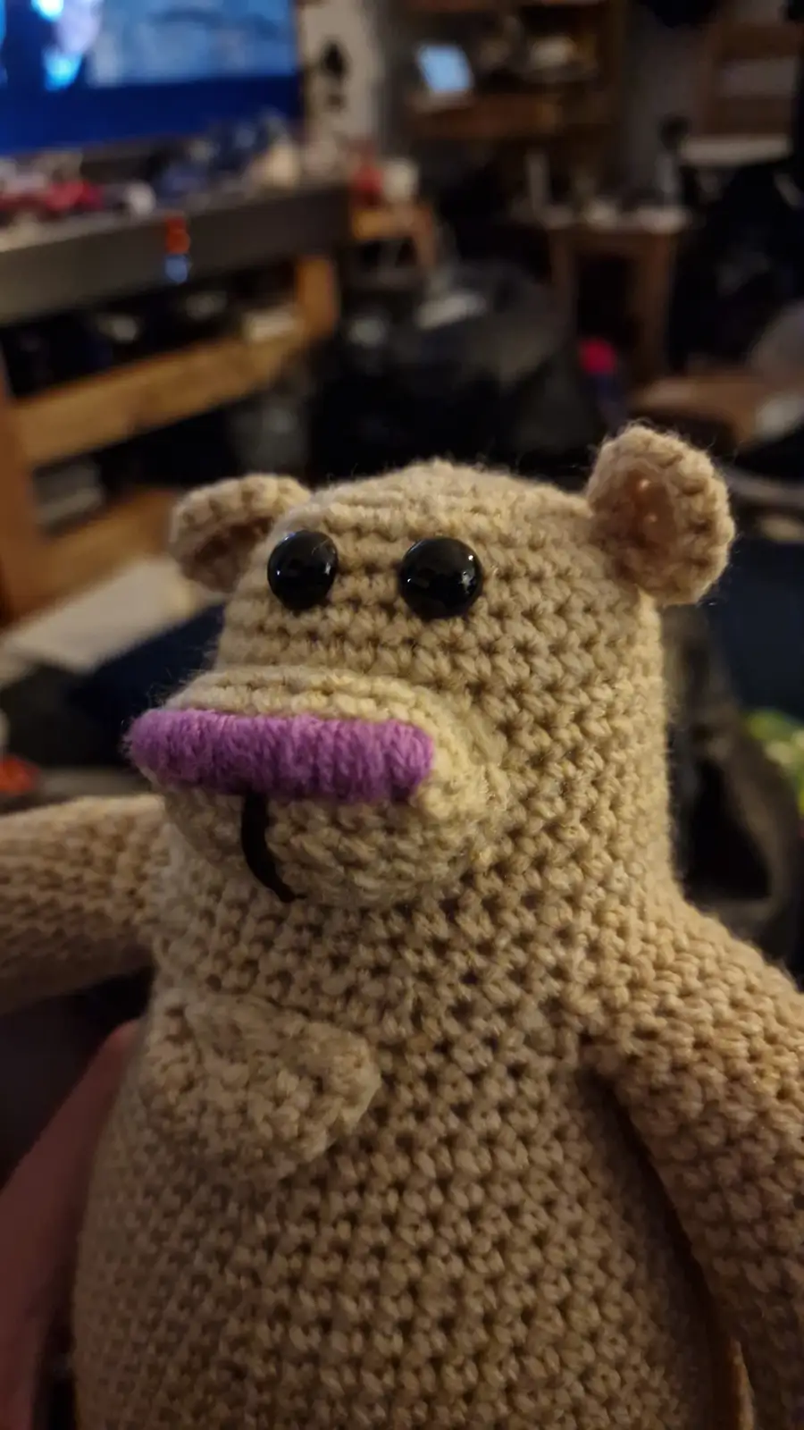 A close-up photo of a beige knitted stuffed animal resembling a bear with large black button eyes, a purple knitted…