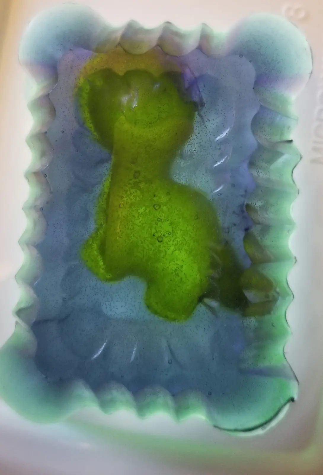 A close-up of a translucent, wavy-edged soap mold filled with colorful soap. The central part is a bright green, roughly…