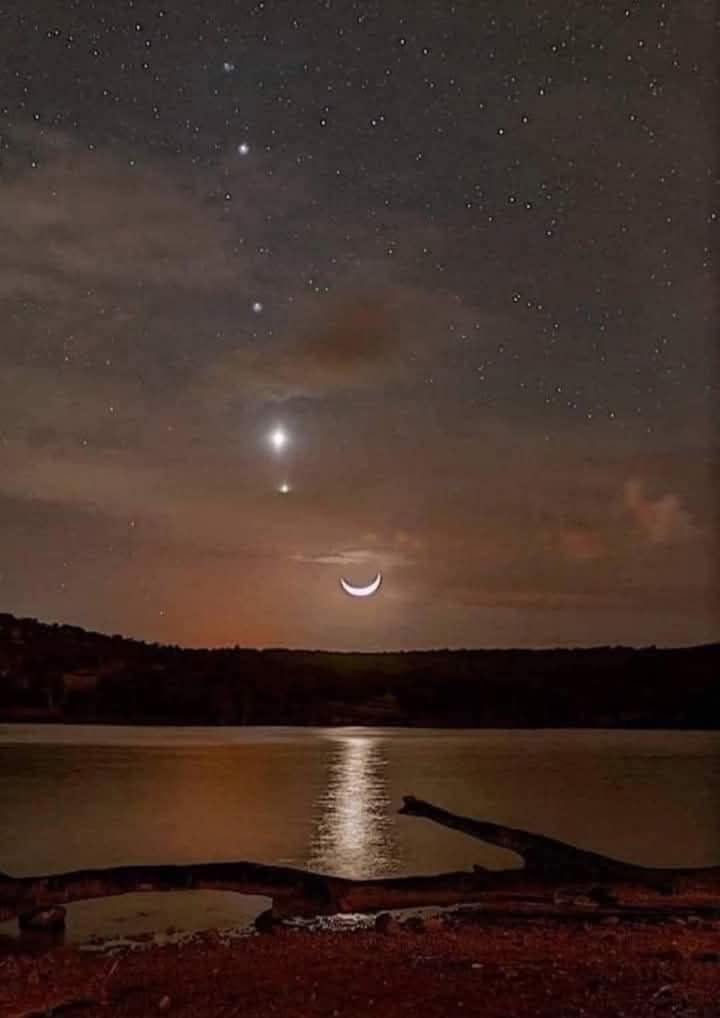 A serene night sky over a calm lake with a crescent moon and several bright stars visible, including a prominent planet or…