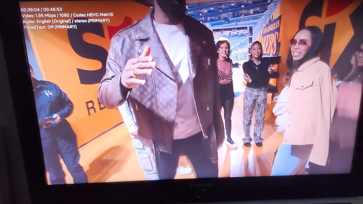 A television screen shows a man in a patterned jacket gesturing while standing on a basketball court with several women…