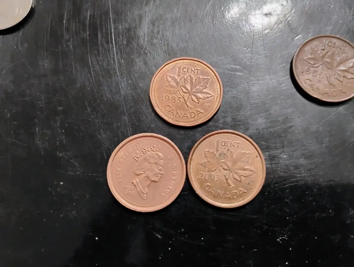 Several Canadian one-cent coins from different years (1959, 1985, 2003) are arranged on a scratched black surface, with one…