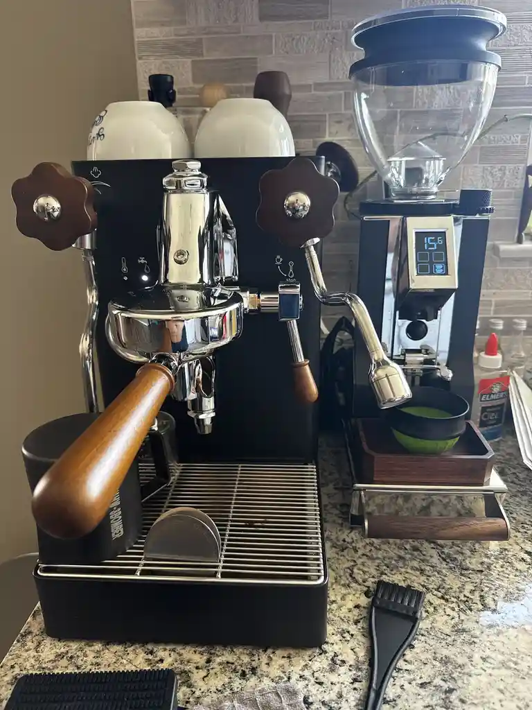 A photo of a high-end espresso machine with wooden handles and chrome parts sits on a granite countertop next to a burr…