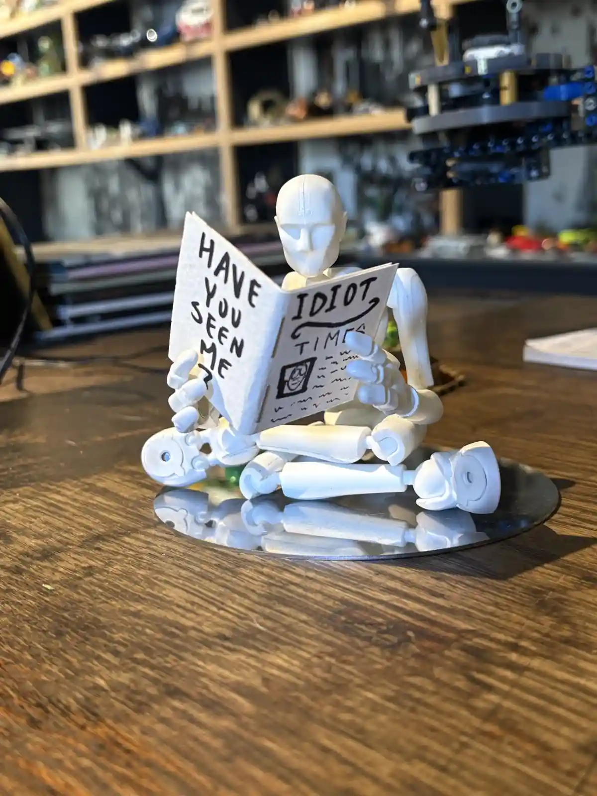 A white, articulated mannequin figure sits cross-legged on a reflective disc atop a wooden table, holding an open paper that…