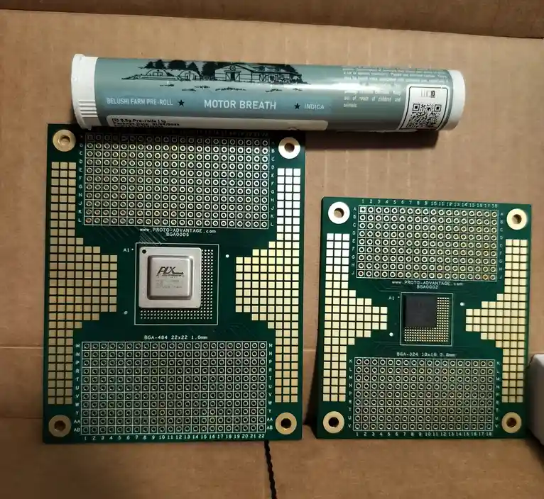 Two green printed circuit boards with BGA chip sockets sit on a cardboard surface, with a cannabis pre-roll tube labeled…
