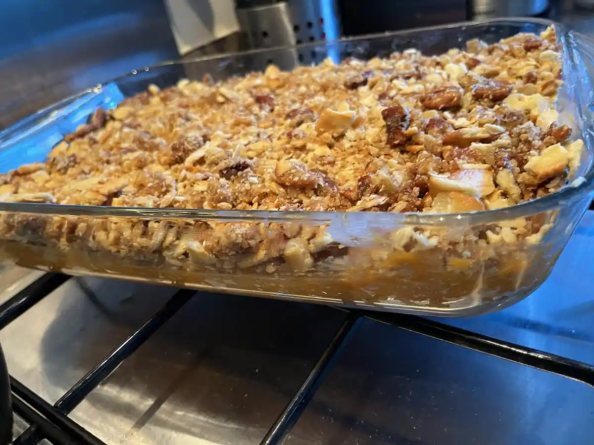 A close-up photo shows a glass baking dish filled with a dessert topped with a golden-brown crumble of oats, nuts, and…