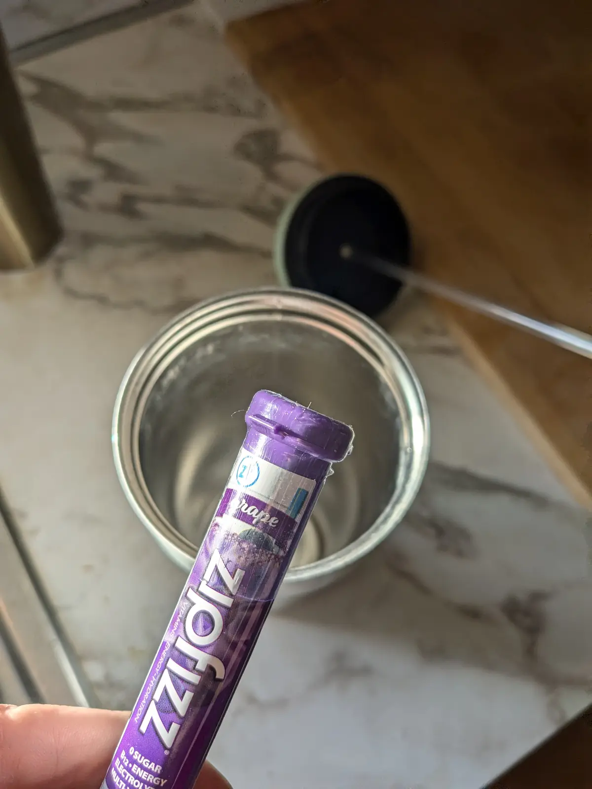A hand holds a purple ZzzFizz grape-flavored energy drink stick above an empty stainless steel travel mug on a marble…