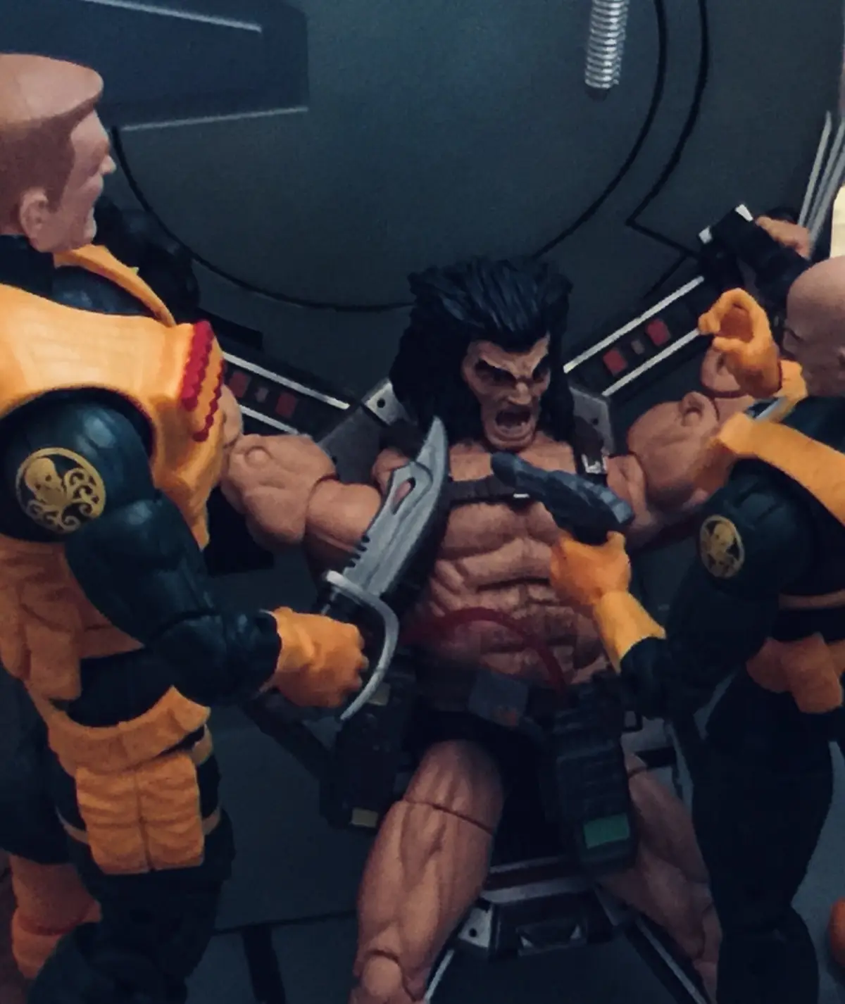 A muscular, shirtless action figure with black hair and an angry expression is restrained in a chair while two other action…