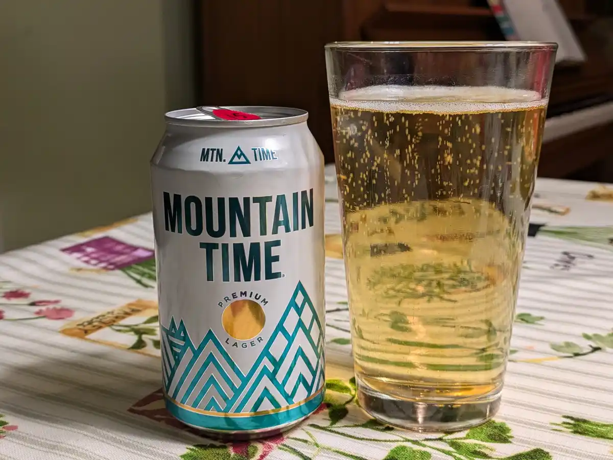 A silver can of 'Mountain Time Premium Lager' sits next to a glass filled with golden beer and rising bubbles, both resting…