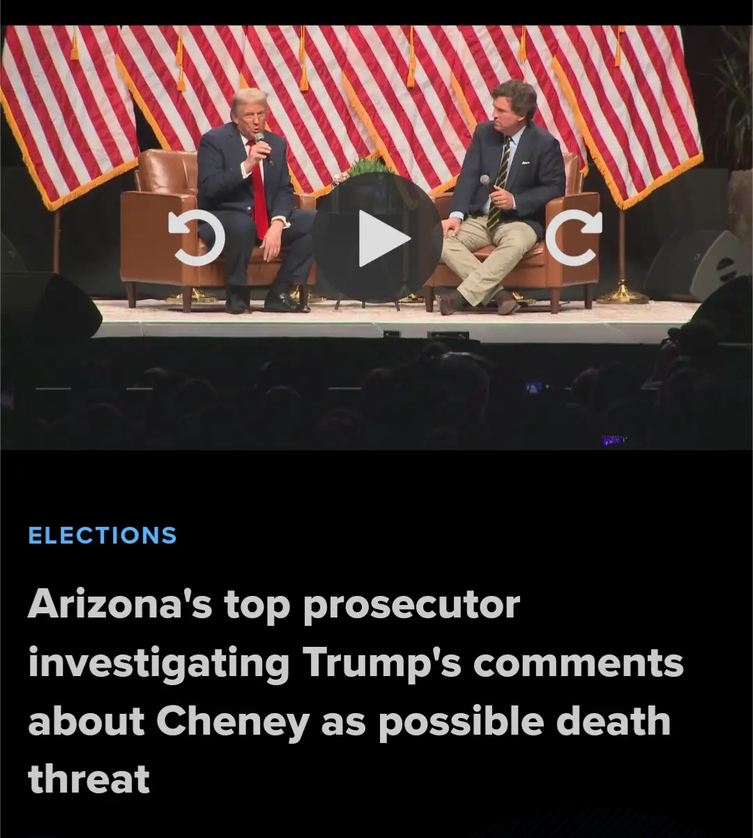 Two men sit in brown armchairs on a stage with red and white striped American flags behind them, overlaid with a video play…
