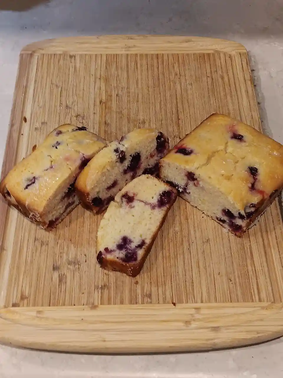 Four slices of a golden-brown berry loaf cake, studded with dark purple berries, are arranged on a light-colored bamboo…