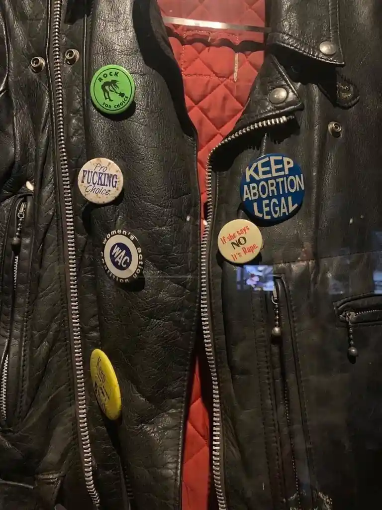 A close-up of a black leather jacket with multiple pro-choice political buttons pinned to it, including 'Rock for Choice'…