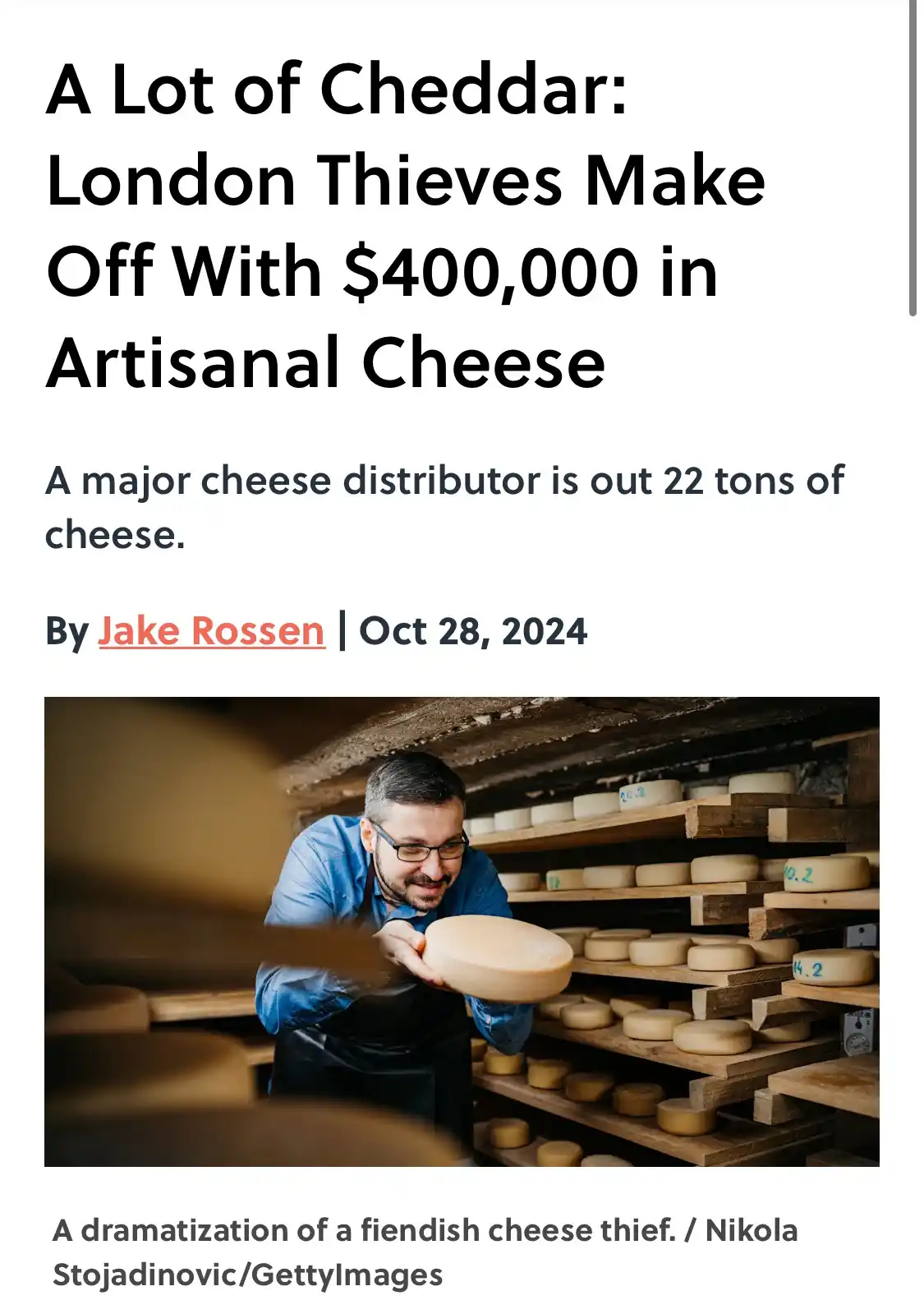 The image shows a man in a blue shirt and black apron, smiling while holding a large round wheel of cheese in a cheese aging…