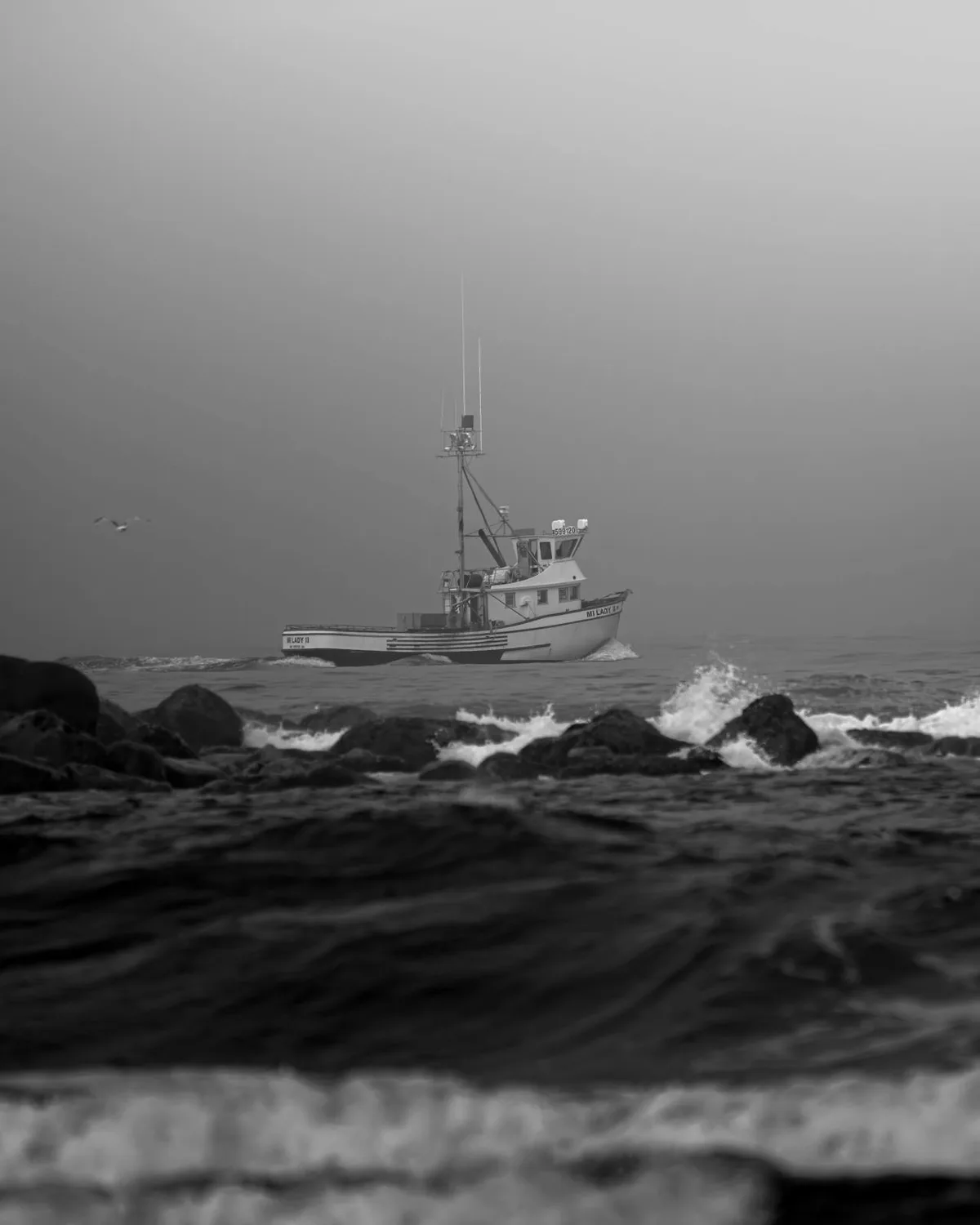A black-and-white photo shows a fishing boat named 'MI LADY II' navigating choppy waters near rocky shores under an overcast…